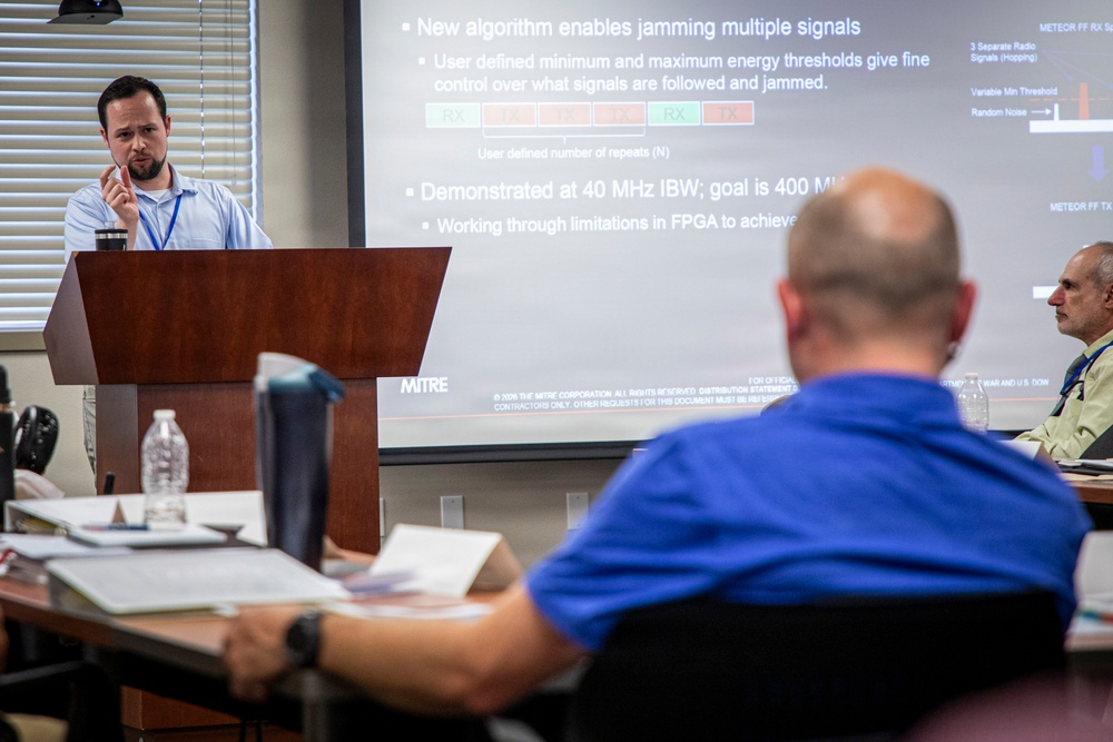 A presenter stands at a wooden podium delivering a technical briefing to an audience in a conference room setting. The projected slide, branded with the MITRE Corporation logo, discusses a new algorithm for jamming multiple signals, referencing FPGA limitations and IBW goals. At least two audience members are visible from behind and to the side, one taking notes.