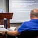 A presenter stands at a wooden podium delivering a technical briefing to an audience in a conference room setting. The projected slide, branded with the MITRE Corporation logo, discusses a new algorithm for jamming multiple signals, referencing FPGA limitations and IBW goals. At least two audience members are visible from behind and to the side, one taking notes.