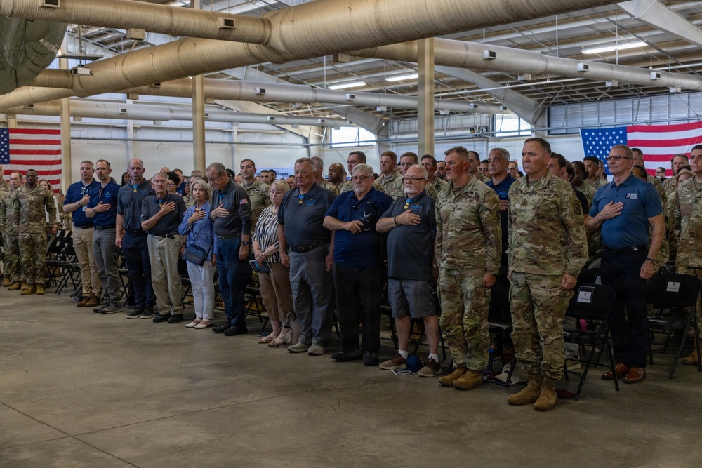A large group of military personnel in camouflage uniforms and civilians stand at attention with hands over their hearts, likely during the Pledge of Allegiance or National Anthem. The event is taking place inside a military hangar or large facility decorated with two large American flags. The gathering appears to be a formal military ceremony or official event.