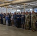 A large group of military personnel in camouflage uniforms and civilians stand at attention with hands over their hearts, likely during the Pledge of Allegiance or National Anthem. The event is taking place inside a military hangar or large facility decorated with two large American flags. The gathering appears to be a formal military ceremony or official event.