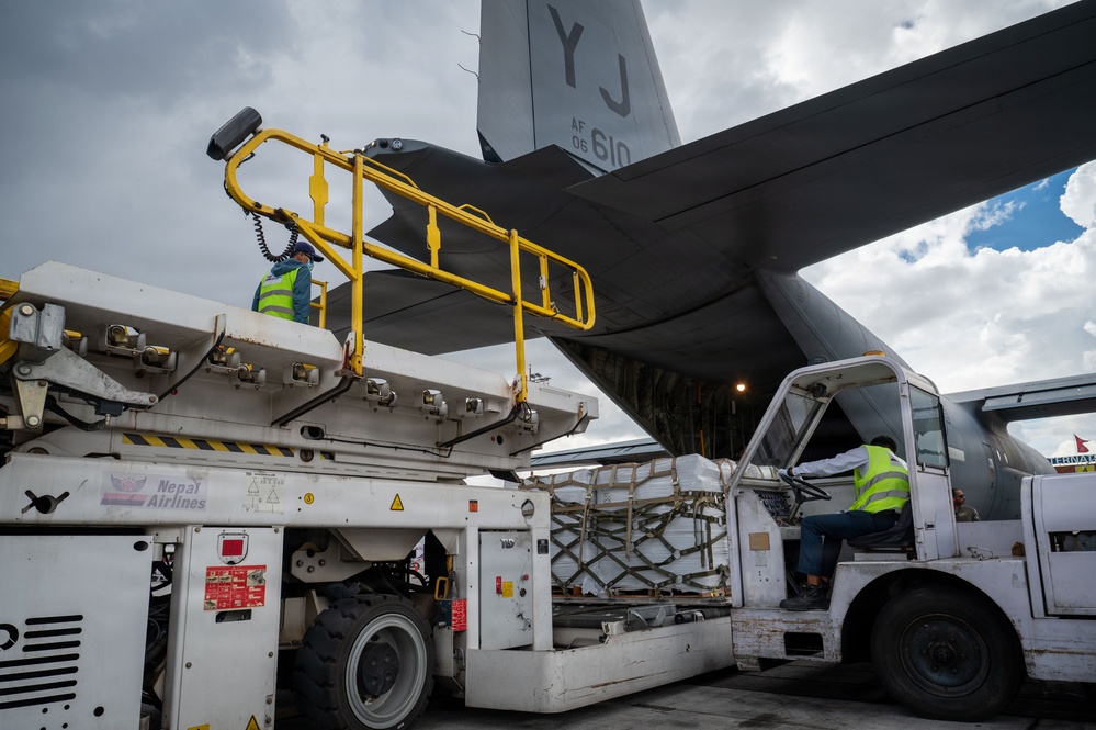 Ground crew workers are loading or unloading cargo from a U.S. Air Force military transport aircraft (tail number AF 06-610, marked 'YJ') at what appears to be an airport in Nepal. A Nepal Airlines ground support vehicle with a cargo loader platform is positioned at the rear ramp of the aircraft. Workers in high-visibility vests are operating the equipment to transfer palletized cargo.