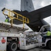 Ground crew workers are loading or unloading cargo from a U.S. Air Force military transport aircraft (tail number AF 06-610, marked 'YJ') at what appears to be an airport in Nepal. A Nepal Airlines ground support vehicle with a cargo loader platform is positioned at the rear ramp of the aircraft. Workers in high-visibility vests are operating the equipment to transfer palletized cargo.