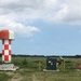 The image shows an aviation navigation or radar facility located in an open field, featuring multiple red-and-white checkered structures characteristic of airport navigation aids. The installation includes what appears to be a rotating radar dome, a large antenna array or ILS (Instrument Landing System) component mounted on a raised platform, and associated equipment cabinets. The facility is situated in a flat, rural landscape consistent with an airport perimeter area.