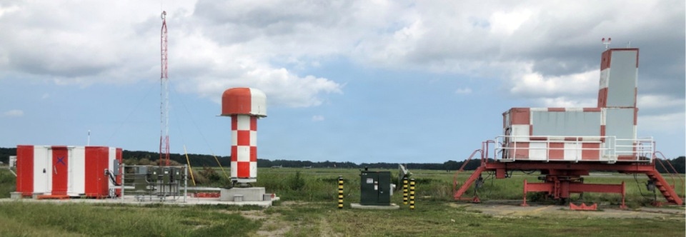 The image shows an aviation navigation or radar facility located in an open field, featuring multiple red-and-white checkered structures characteristic of airport navigation aids. The installation includes what appears to be a rotating radar dome, a large antenna array or ILS (Instrument Landing System) component mounted on a raised platform, and associated equipment cabinets. The facility is situated in a flat, rural landscape consistent with an airport perimeter area.
