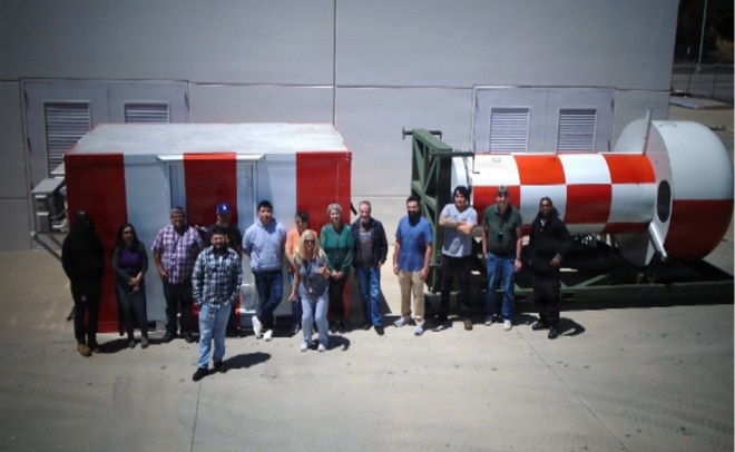 A group of approximately 14 people are posing outdoors in front of two large pieces of equipment painted with distinctive red and white markings. The equipment appears to be industrial or aerospace-related, including what looks like a cylindrical component with a red and white checkered pattern on the right. The setting appears to be an industrial or aerospace facility with a large gray building in the background.