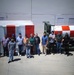 A group of approximately 14 people are posing outdoors in front of two large pieces of equipment painted with distinctive red and white markings. The equipment appears to be industrial or aerospace-related, including what looks like a cylindrical component with a red and white checkered pattern on the right. The setting appears to be an industrial or aerospace facility with a large gray building in the background.