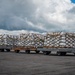The image shows multiple flatbed cargo dollies loaded with palletized freight secured with cargo nets and strapping on an airport tarmac. The cargo consists of white boxes/packages stacked on standard air freight pallets, suggesting recent unloading from or preparation for loading onto an aircraft. Mountains and a partially cloudy sky are visible in the background, indicating a location near elevated terrain.