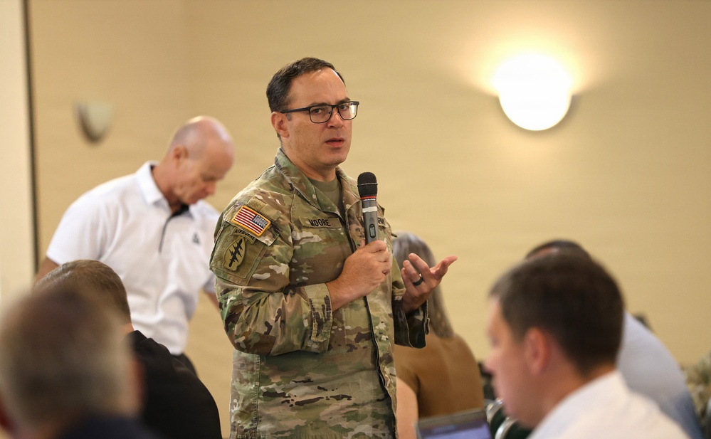 A U.S. Army soldier in MultiCam (OCP) uniform is speaking into a handheld microphone, apparently addressing an audience at an indoor event or briefing. The soldier's name tape reads 'MOORE' and patches include an American flag, an Airborne tab, and a Special Forces patch. Several civilians and other attendees are visible in the foreground and background.