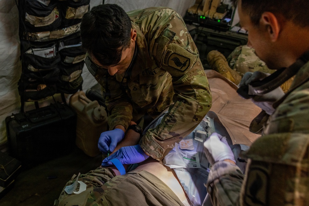 Two U.S. Army soldiers in OCP (Operational Camouflage Pattern) uniforms are providing medical treatment to a casualty lying on a surface. The primary medic, wearing blue nitrile gloves, appears to be performing an intravenous (IV) insertion or wound care on the patient's arm/wrist area. The setting appears to be a field medical station or aid tent with medical supplies visible.