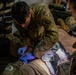 Two U.S. Army soldiers in OCP (Operational Camouflage Pattern) uniforms are providing medical treatment to a casualty lying on a surface. The primary medic, wearing blue nitrile gloves, appears to be performing an intravenous (IV) insertion or wound care on the patient's arm/wrist area. The setting appears to be a field medical station or aid tent with medical supplies visible.