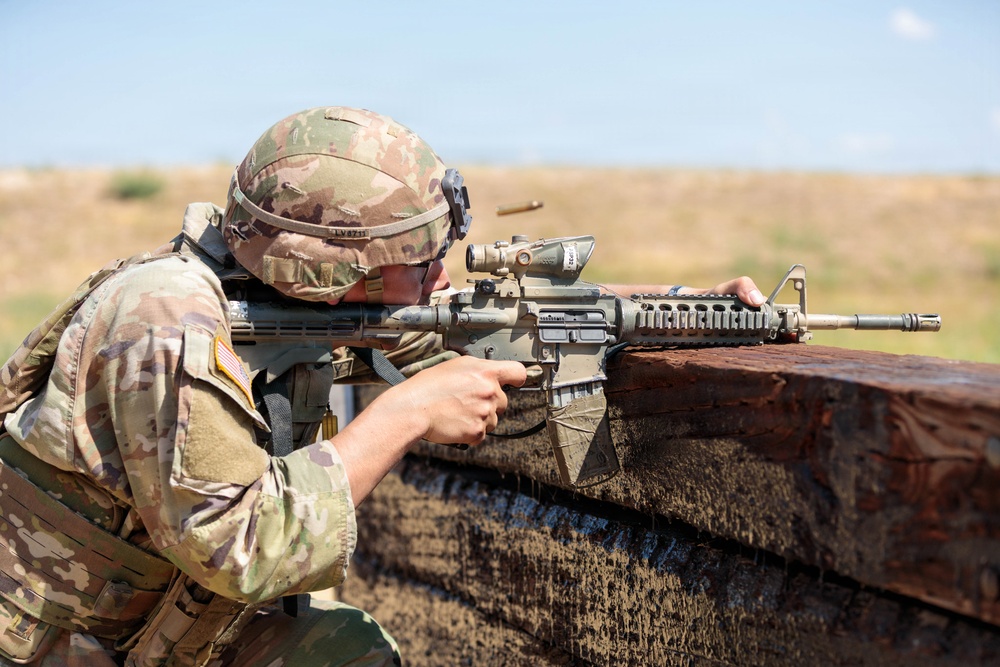 A U.S. Army soldier in full combat gear is firing a rifle from behind a fortified barrier, likely during a training exercise or live-fire range event. A spent brass casing is visibly ejecting from the weapon. The setting appears to be an arid, open training range under clear skies.