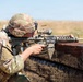 A U.S. Army soldier in full combat gear is firing a rifle from behind a fortified barrier, likely during a training exercise or live-fire range event. A spent brass casing is visibly ejecting from the weapon. The setting appears to be an arid, open training range under clear skies.