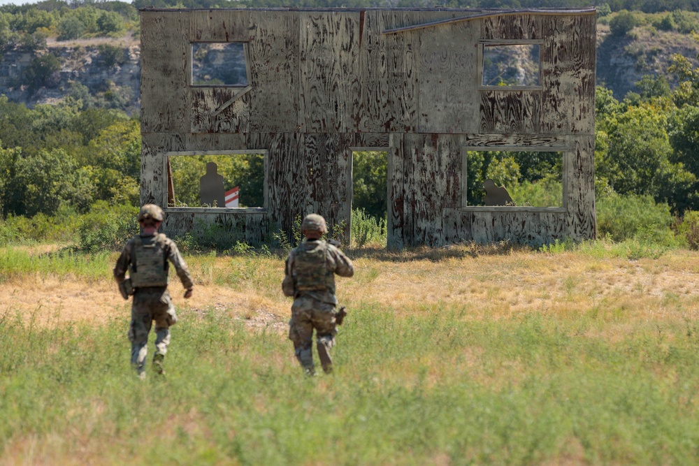 Two soldiers in camouflage uniforms and tactical gear are approaching a weathered, two-story plywood training structure in an open field. The structure appears to be a military training facility mock building used for tactical exercises, with visible silhouette targets inside. The setting is a rural, vegetated landscape consistent with a military training range.