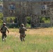 Two soldiers in camouflage uniforms and tactical gear are approaching a weathered, two-story plywood training structure in an open field. The structure appears to be a military training facility mock building used for tactical exercises, with visible silhouette targets inside. The setting is a rural, vegetated landscape consistent with a military training range.