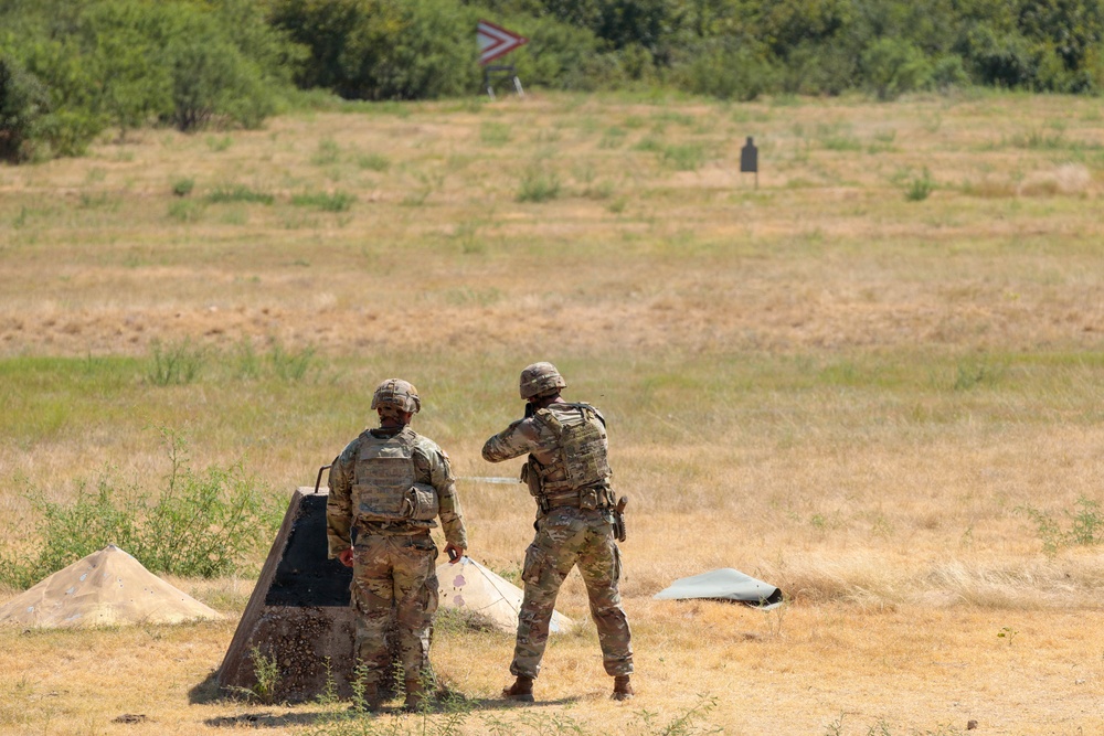 Two soldiers in camouflage uniforms and tactical gear are positioned at what appears to be a military firing range. They are facing downrange toward a silhouette target visible in the distance. The setting is an open grassy field with trees in the background and range infrastructure visible.