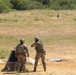 Two soldiers in camouflage uniforms and tactical gear are positioned at what appears to be a military firing range. They are facing downrange toward a silhouette target visible in the distance. The setting is an open grassy field with trees in the background and range infrastructure visible.