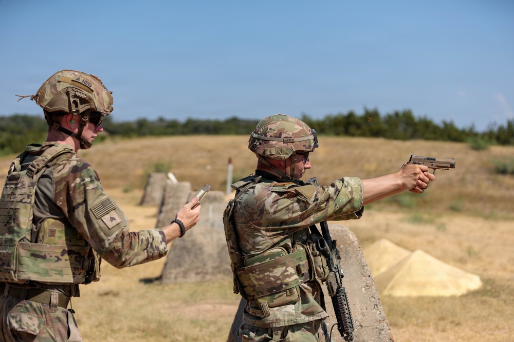 Two military personnel in full combat gear are on an outdoor shooting range. One soldier is actively firing a pistol in a two-handed stance while the other observes and appears to be scoring or recording results on a card. The setting is a dry, open-air range with concrete barriers and target markers visible downrange.