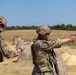 Two military personnel in full combat gear are on an outdoor shooting range. One soldier is actively firing a pistol in a two-handed stance while the other observes and appears to be scoring or recording results on a card. The setting is a dry, open-air range with concrete barriers and target markers visible downrange.