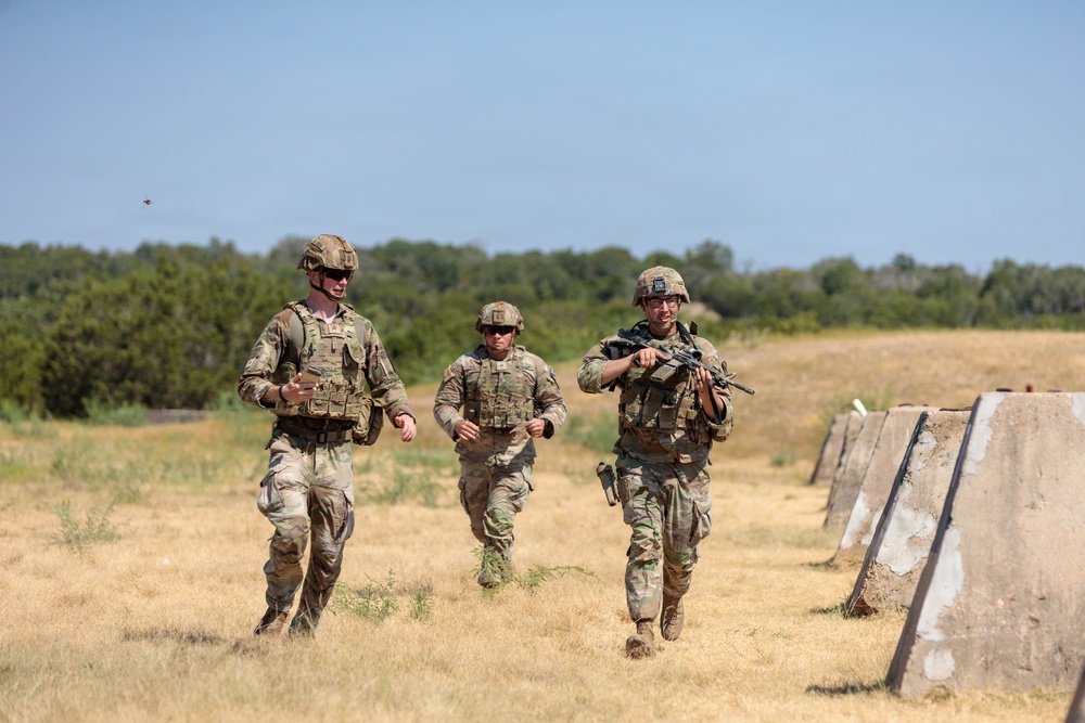 Three military personnel in full combat gear are jogging across an open grassy field during what appears to be a training exercise. They are wearing matching OCP (Operational Camouflage Pattern) uniforms with plate carriers and helmets. Concrete barriers are visible along the right side of the frame, consistent with a military training range environment.