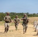 Three military personnel in full combat gear are jogging across an open grassy field during what appears to be a training exercise. They are wearing matching OCP (Operational Camouflage Pattern) uniforms with plate carriers and helmets. Concrete barriers are visible along the right side of the frame, consistent with a military training range environment.