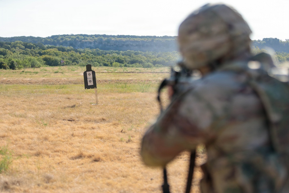 A military or law enforcement personnel in full camouflage gear and helmet is aiming a rifle at a standard silhouette paper target on an outdoor shooting range. The shooter is positioned in the foreground, slightly out of focus, while the target is clearly visible in the mid-ground against a backdrop of dry grassland and tree-lined hills. Smoke is faintly visible near the shooter, suggesting a recent or ongoing discharge.