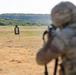 A military or law enforcement personnel in full camouflage gear and helmet is aiming a rifle at a standard silhouette paper target on an outdoor shooting range. The shooter is positioned in the foreground, slightly out of focus, while the target is clearly visible in the mid-ground against a backdrop of dry grassland and tree-lined hills. Smoke is faintly visible near the shooter, suggesting a recent or ongoing discharge.