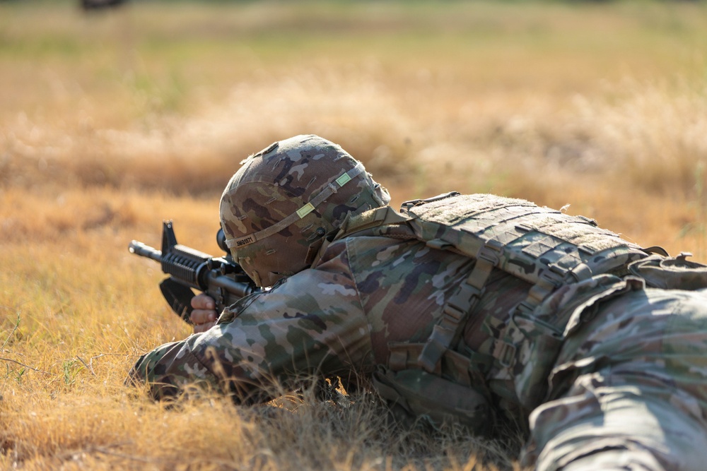 A soldier in full combat gear is lying prone in a dry grass field, aiming a rifle downrange. The individual is wearing MultiCam/OCP camouflage uniform, a combat helmet, and a plate carrier vest. The image appears to depict a military marksmanship training exercise.