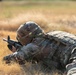 A soldier in full combat gear is lying prone in a dry grass field, aiming a rifle downrange. The individual is wearing MultiCam/OCP camouflage uniform, a combat helmet, and a plate carrier vest. The image appears to depict a military marksmanship training exercise.