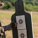 A military service member in full camouflage uniform and tactical gear is examining a shooting target at an outdoor firing range. The target consists of a human-silhouette backer board with two paper bullseye targets attached, the upper one showing multiple bullet impact holes concentrated near the center. The setting appears to be a daytime training exercise in an open field environment.