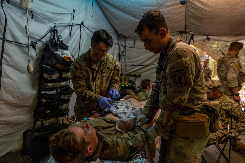 Two U.S. Army soldiers in OCP (Operational Camouflage Pattern) uniforms are providing medical treatment to a casualty lying on a field litter inside a military medical tent. One medic is wearing blue nitrile gloves and appears to be applying or adjusting bandaging/dressings on the patient. The scene appears to be a field medical training exercise or combat casualty care scenario.