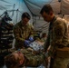 Two U.S. Army soldiers in OCP (Operational Camouflage Pattern) uniforms are providing medical treatment to a casualty lying on a field litter inside a military medical tent. One medic is wearing blue nitrile gloves and appears to be applying or adjusting bandaging/dressings on the patient. The scene appears to be a field medical training exercise or combat casualty care scenario.