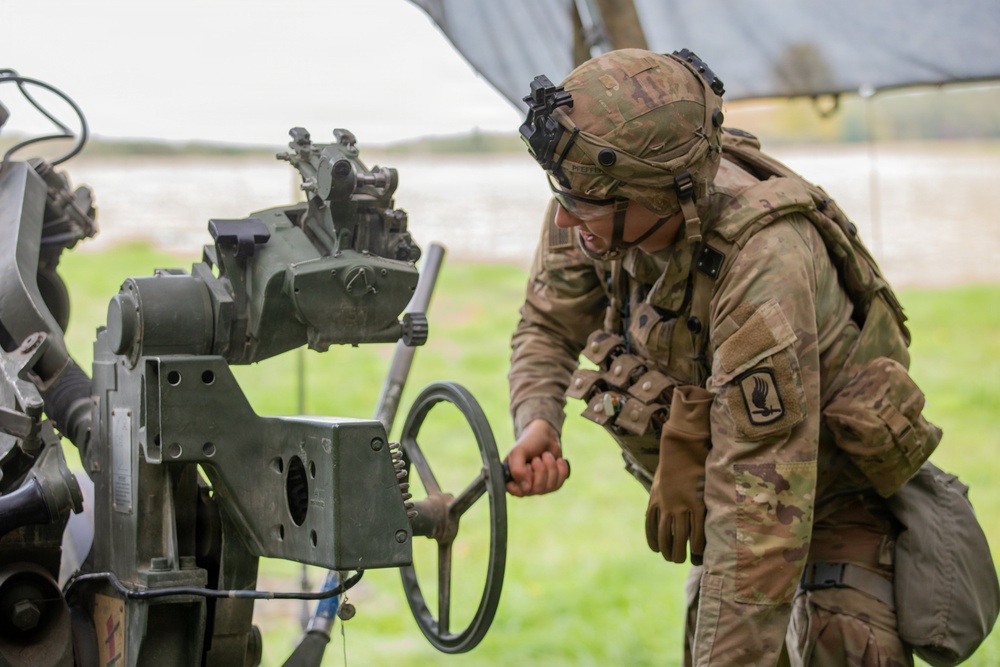 A U.S. Army soldier in full combat gear is operating or adjusting what appears to be an artillery piece, likely an M777 howitzer or similar field artillery system. The soldier is wearing Operational Camouflage Pattern (OCP) uniform with a unit patch visible on the left shoulder, a combat helmet with night vision mount, and tactical vest. The activity appears to be taking place at an outdoor training or operational field site.