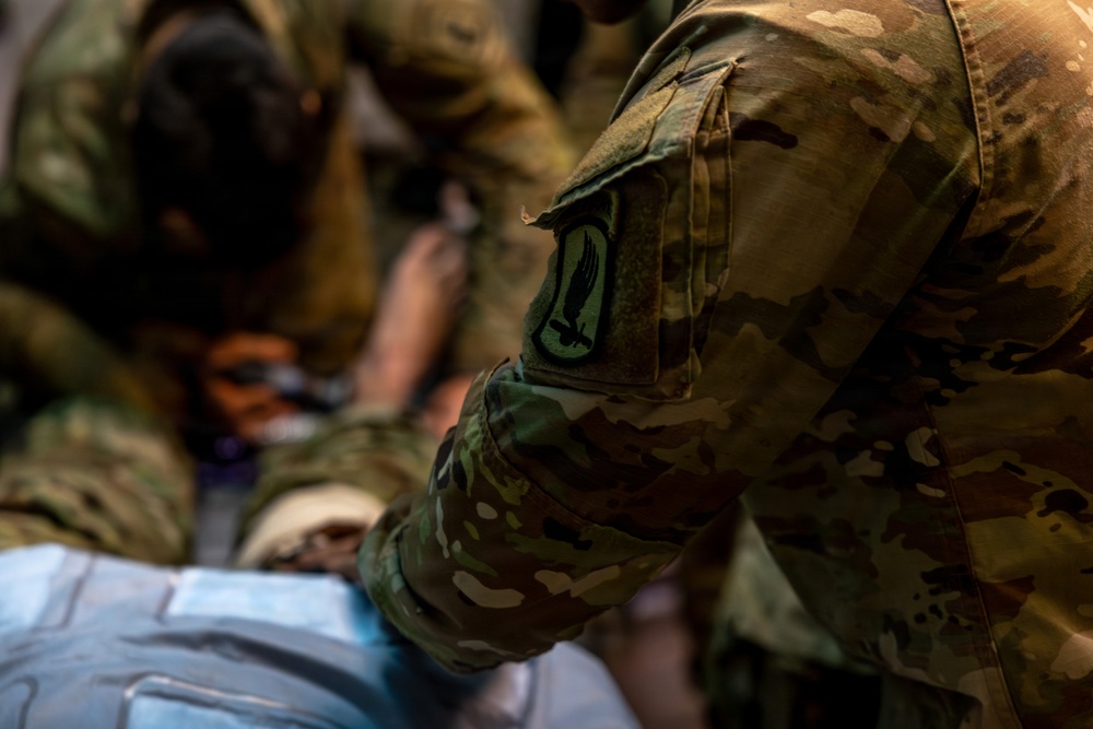 The image shows military personnel in OCP (Operational Camouflage Pattern) uniforms attending to a person lying down, likely during a medical training exercise or casualty care scenario. A shoulder patch featuring what appears to be a dagger or sword emblem is clearly visible on the foreground soldier's left sleeve. Multiple personnel are working together in close proximity over the subject.