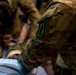 The image shows military personnel in OCP (Operational Camouflage Pattern) uniforms attending to a person lying down, likely during a medical training exercise or casualty care scenario. A shoulder patch featuring what appears to be a dagger or sword emblem is clearly visible on the foreground soldier's left sleeve. Multiple personnel are working together in close proximity over the subject.