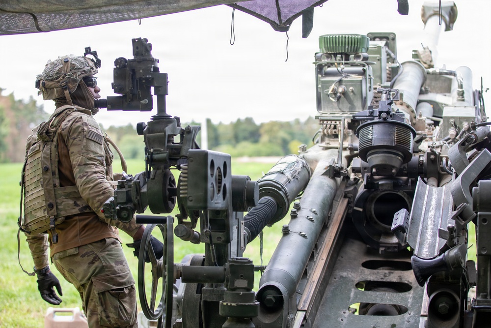 A soldier in full combat gear and camouflage uniform is operating or inspecting a large artillery piece, likely an M777 howitzer or similar towed artillery system. The soldier is wearing a helmet with mounted optics, body armor with MOLLE webbing, and black tactical gloves. The setting appears to be an outdoor training or operational field environment with trees visible in the background.