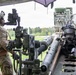 A soldier in full combat gear and camouflage uniform is operating or inspecting a large artillery piece, likely an M777 howitzer or similar towed artillery system. The soldier is wearing a helmet with mounted optics, body armor with MOLLE webbing, and black tactical gloves. The setting appears to be an outdoor training or operational field environment with trees visible in the background.