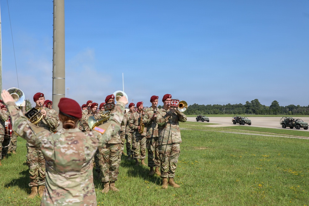 A U.S. Army military band wearing distinctive maroon berets performs outdoors on a grassy area adjacent to a paved airfield or parade ground. A conductor with raised arms leads the ensemble, which includes brass and woodwind instruments. Military utility vehicles are visible in the background on the tarmac, suggesting a formal ceremony or welcome event.