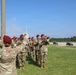 A U.S. Army military band wearing distinctive maroon berets performs outdoors on a grassy area adjacent to a paved airfield or parade ground. A conductor with raised arms leads the ensemble, which includes brass and woodwind instruments. Military utility vehicles are visible in the background on the tarmac, suggesting a formal ceremony or welcome event.