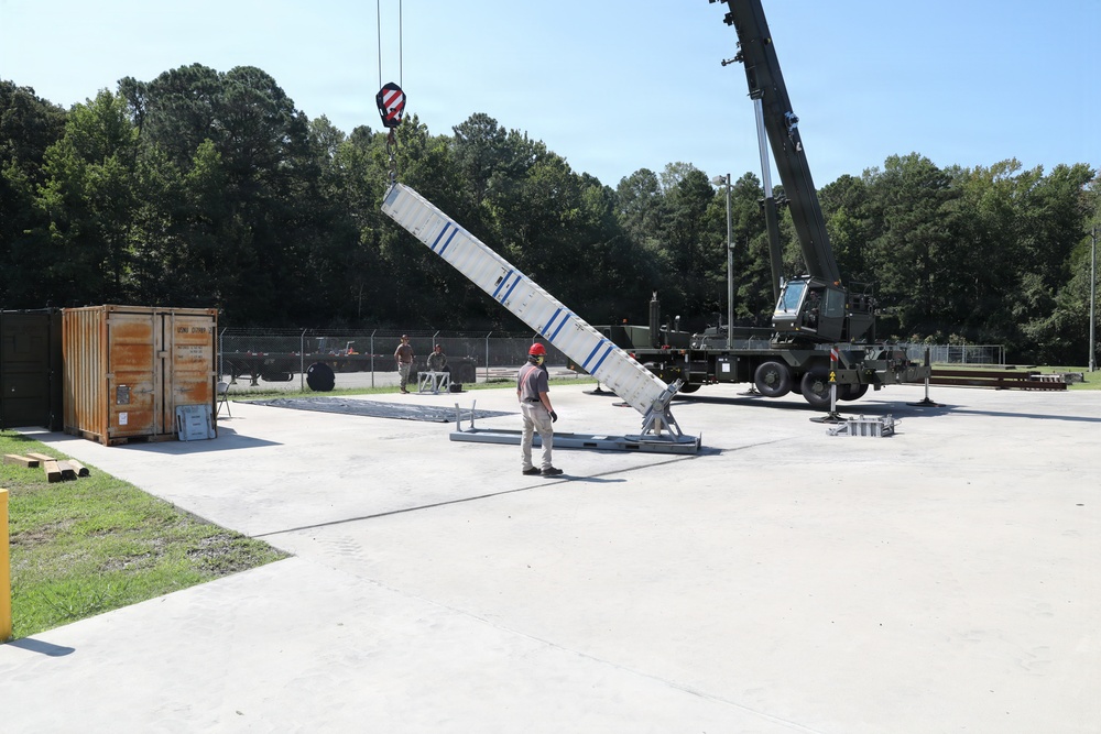 A military or industrial crane operation is underway on a concrete pad, where a large white elongated structure with blue markings is being lifted or positioned by a mobile crane. A worker wearing a red hard hat is standing nearby, appearing to guide or supervise the lift. The setting appears to be a military installation or training facility, surrounded by a chain-link fence and wooded area.