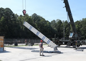 A military or industrial crane operation is underway on a concrete pad, where a large white elongated structure with blue markings is being lifted or positioned by a mobile crane. A worker wearing a red hard hat is standing nearby, appearing to guide or supervise the lift. The setting appears to be a military installation or training facility, surrounded by a chain-link fence and wooded area.