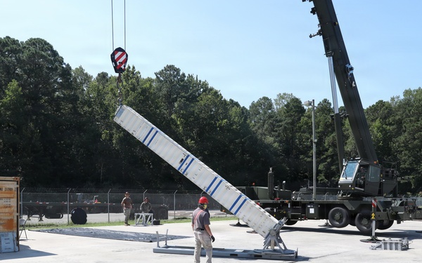 A military or industrial crane operation is underway on a concrete pad, where a large white elongated structure with blue markings is being lifted or positioned by a mobile crane. A worker wearing a red hard hat is standing nearby, appearing to guide or supervise the lift. The setting appears to be a military installation or training facility, surrounded by a chain-link fence and wooded area.