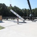 A military or industrial crane operation is underway on a concrete pad, where a large white elongated structure with blue markings is being lifted or positioned by a mobile crane. A worker wearing a red hard hat is standing nearby, appearing to guide or supervise the lift. The setting appears to be a military installation or training facility, surrounded by a chain-link fence and wooded area.
