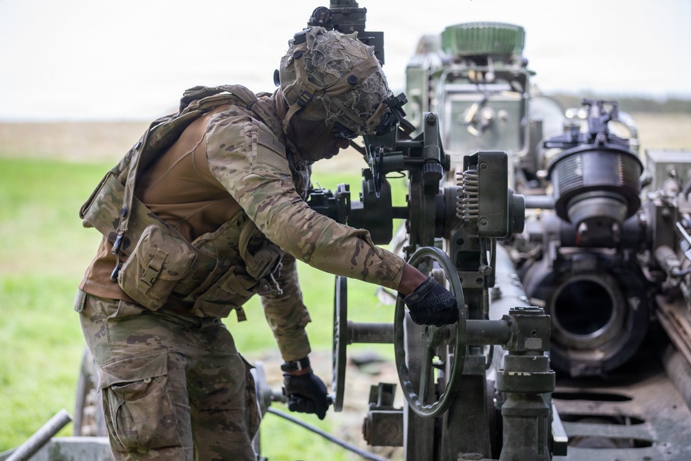 A soldier in full camouflage combat gear is operating or adjusting the elevation/traverse mechanism of what appears to be an artillery piece, likely an M777 howitzer or similar towed artillery system. The soldier is wearing a multicam uniform, tactical vest, black gloves, and a camouflage-netted helmet with night vision mount. The setting appears to be an open field training environment.