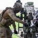 A soldier in full camouflage combat gear is operating or adjusting the elevation/traverse mechanism of what appears to be an artillery piece, likely an M777 howitzer or similar towed artillery system. The soldier is wearing a multicam uniform, tactical vest, black gloves, and a camouflage-netted helmet with night vision mount. The setting appears to be an open field training environment.