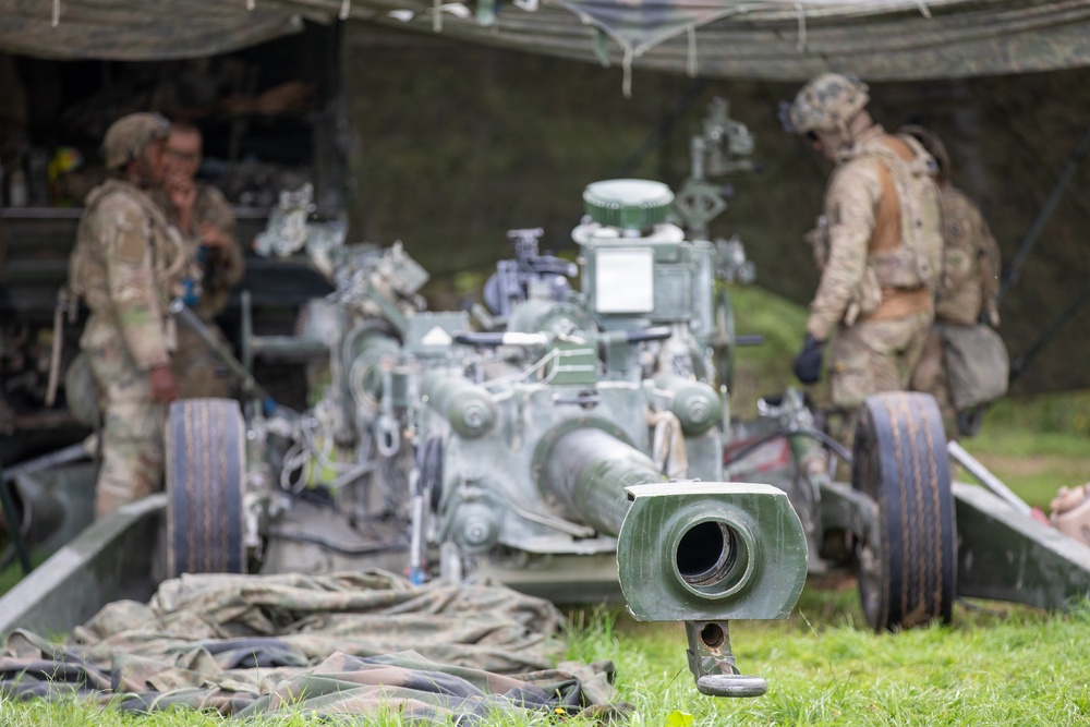 The image shows military personnel in camouflage uniforms working around what appears to be a towed artillery piece, likely an M777 howitzer or similar field gun, set up under a camouflage netting canopy. In the foreground, a muzzle brake or gun component rests on the grass, sharply in focus while the background elements are blurred. The scene depicts an active field artillery position during what appears to be a training exercise or operational deployment.