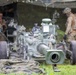The image shows military personnel in camouflage uniforms working around what appears to be a towed artillery piece, likely an M777 howitzer or similar field gun, set up under a camouflage netting canopy. In the foreground, a muzzle brake or gun component rests on the grass, sharply in focus while the background elements are blurred. The scene depicts an active field artillery position during what appears to be a training exercise or operational deployment.