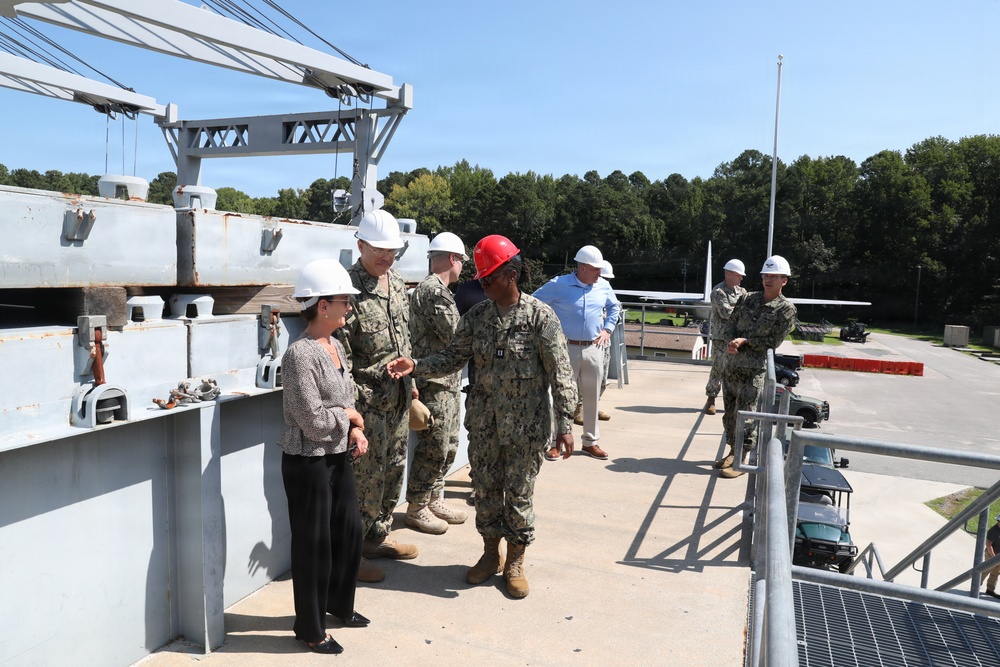 A group of military personnel in Navy camouflage uniforms and civilians are conducting a facility tour or inspection at what appears to be a naval installation or shipyard. All individuals are wearing hard hats, with most wearing white hard hats and one person wearing a red hard hat. A large crane structure and what appears to be a vessel or dock structure are visible in the background.