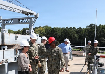 A group of military personnel in Navy camouflage uniforms and civilians are conducting a facility tour or inspection at what appears to be a naval installation or shipyard. All individuals are wearing hard hats, with most wearing white hard hats and one person wearing a red hard hat. A large crane structure and what appears to be a vessel or dock structure are visible in the background.