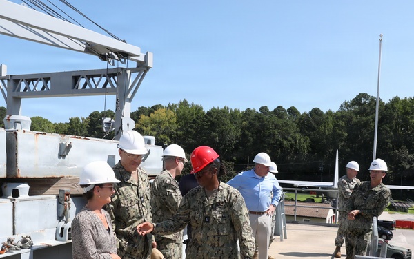 A group of military personnel in Navy camouflage uniforms and civilians are conducting a facility tour or inspection at what appears to be a naval installation or shipyard. All individuals are wearing hard hats, with most wearing white hard hats and one person wearing a red hard hat. A large crane structure and what appears to be a vessel or dock structure are visible in the background.