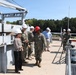 A group of military personnel in Navy camouflage uniforms and civilians are conducting a facility tour or inspection at what appears to be a naval installation or shipyard. All individuals are wearing hard hats, with most wearing white hard hats and one person wearing a red hard hat. A large crane structure and what appears to be a vessel or dock structure are visible in the background.
